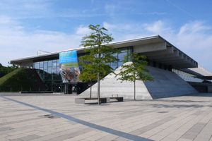 Zu sehen ist die Glasfront der Biosphäre Potsdam am Volkspark, im Vordergrund ist eine steinerne Fläche mit ein paar Bäumen.