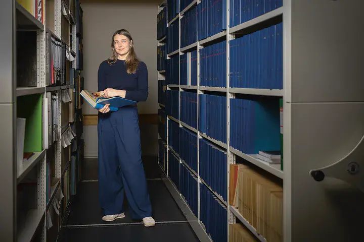 Dr. Elisabeth Kimmerle, Wissenschaftliche Mitarbeiterin Leibniz- Zentrum für Zeithistorische Forschung Eine junge Frau steht in einer Bibliothek und hält ein Buch aufgeklappt in der Hand