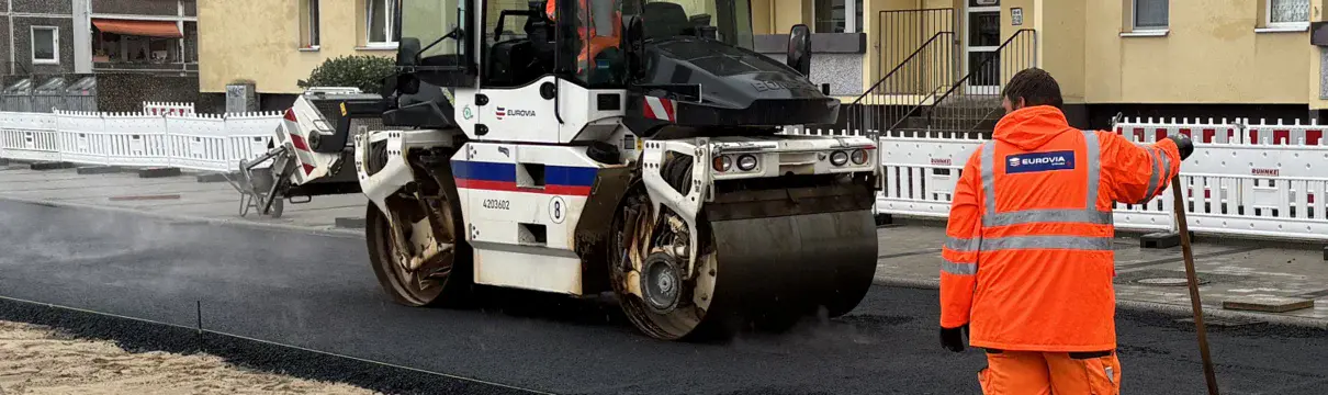 Orange gekleideter Bauarbeiter und ein Baufahrzeug an einer Straße.