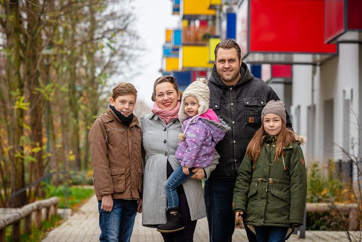 Familie Drygalla, bestehend aus Vater, Mutter und drei Kindern blickt in die Kamera. Im Hintergrund erstreckt sich die Konrad-Wolf-Allee.