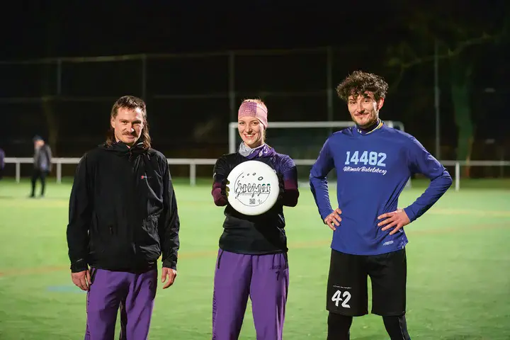 Drei junge Menschen stehen nebeneinander auf dem Rasen eines Sportplatzes. Die Frau in der Mitte hält eine Frisbee-Scheibe hoch.