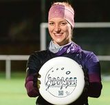 Drei junge Menschen stehen nebeneinander auf dem Rasen eines Sportplatzes. Die Frau in der Mitte hält eine Frisbee-Scheibe hoch.