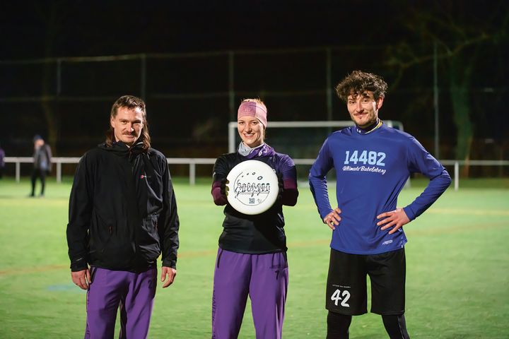 Drei junge Menschen stehen nebeneinander auf dem Rasen eines Sportplatzes. Die Frau in der Mitte hält eine Frisbee-Scheibe hoch.