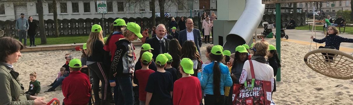 Eine Gruppe von Kindern mit grünen Cappies stehen auf einem Spielplatz. Im Hintergrund stehen der Geschäftsführer der ProPotsdam und der Baubeigeordnete der Stadt.