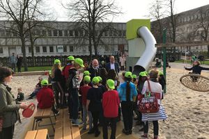 Eine Gruppe von Kindern mit grünen Cappies stehen auf einem Spielplatz. Im Hintergrund stehen der Geschäftsführer der ProPotsdam und der Baubeigeordnete der Stadt.
