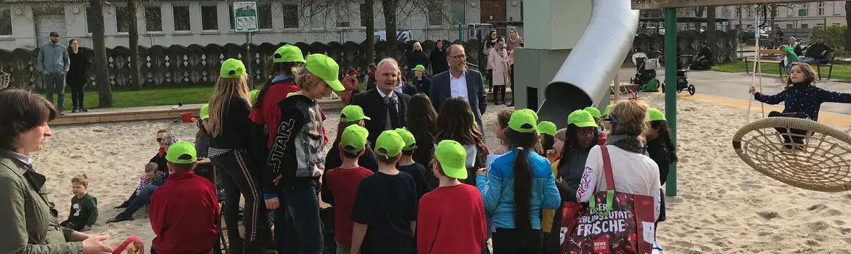 Einweihung Plantage Eine Gruppe von Kindern mit grünen Cappies stehen auf einem Spielplatz. Im Hintergrund stehen der Geschäftsführer der ProPotsdam und der Baubeigeordnete der Stadt.