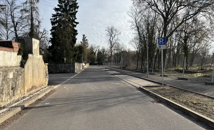 Leere Straße mit Parkstreifen und Parkschild, links eine Mauer und rechts ein eingezäuntes Gelände mit Bäumen.