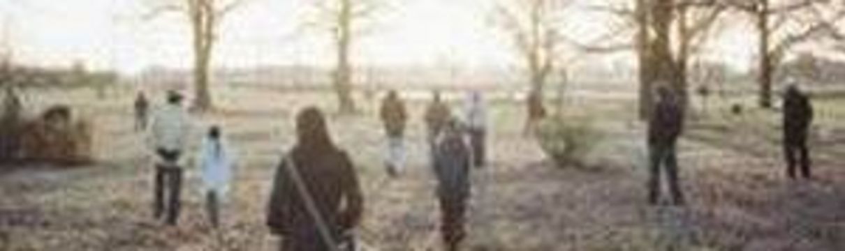 Menschen in Winterjacken stehen auf einer parkähnlichen Rasenfläche. Im Hintergrund scheint eine winterliche Sonne.