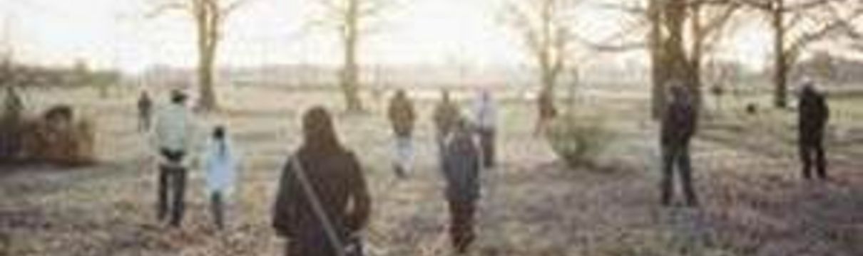 Menschen in Winterjacken stehen auf einer parkähnlichen Rasenfläche. Im Hintergrund scheint eine winterliche Sonne.