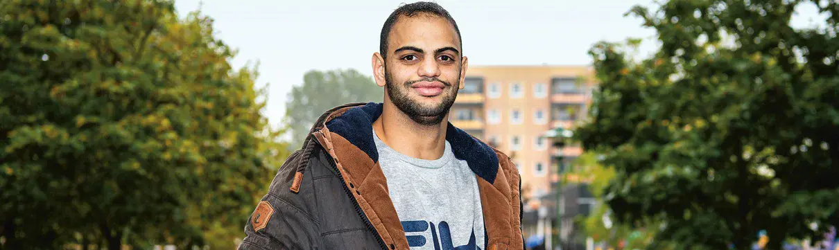 Junger, freundlicher Mann in grauem Pullover und grau-brauner Jacke steht auf einem betoniertem Weg, der zu einem Plattenbau führt. 