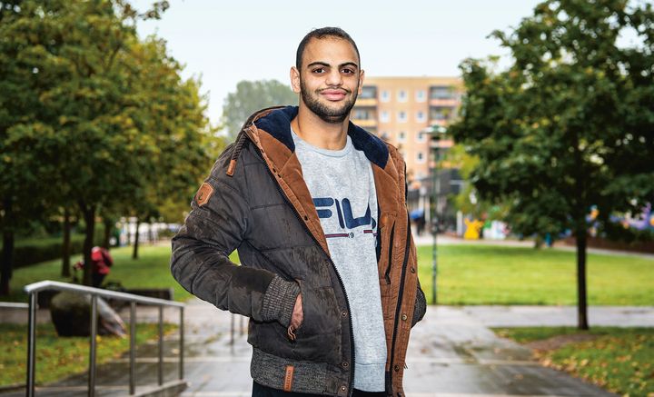Junger, freundlicher Mann in grauem Pullover und grau-brauner Jacke steht auf einem betoniertem Weg, der zu einem Plattenbau führt. 