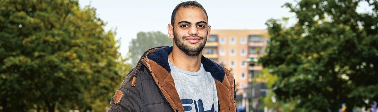 Junger, freundlicher Mann in grauem Pullover und grau-brauner Jacke steht auf einem betoniertem Weg, der zu einem Plattenbau führt. 