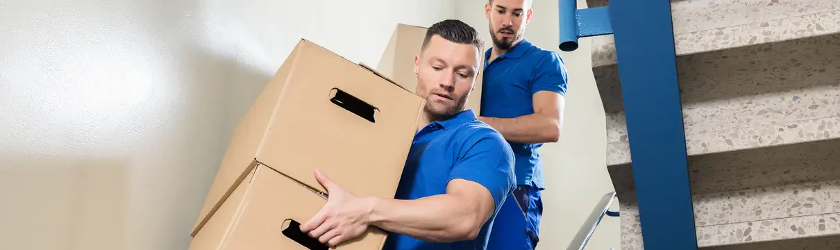 Treppenhaus, in dem zwei Männer in blauer Arbeitskleidung Umzugkartons von oben nach unten tragen