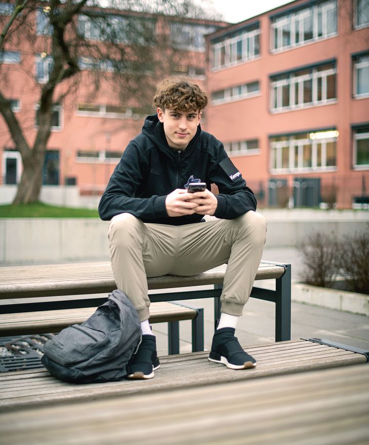 Ein 18-jähriger Mann mit blauer Kapuzenjacke und Mobiltelefon vor einem Gebäude