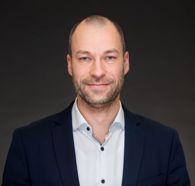 Porträtbild Mann mit kurzem Bart, hellem Hemd und blauem Sacko vor dunklem Hintergrund