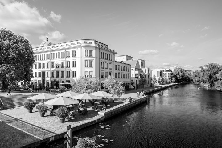 Schwarz-Weiß-Aufnahme einer Uferpromenade, in deren Vordergrund Gastronomiebetrieb unter Sonnenschirmen sichtbar ist