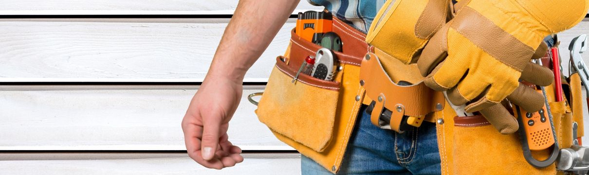 Closeup Werkzeuggürtel mit verschiedenen Werkzeugen an Handwerker in Jeans und Karohemd mit Arbeitshandschuh an der einen Hand, die auch einen gelben Bauhelm hält.