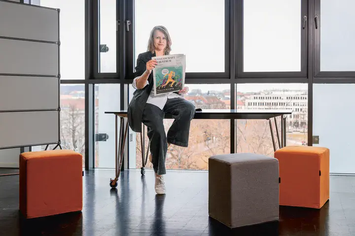 Dr. Simone Leinkauf, Geschäftsführerin des Vereins proWissen Potsdam Eine Frau sitzt auf einem Tisch und schaut den Betrachter an, in der Hand hält sie eine Zeitung