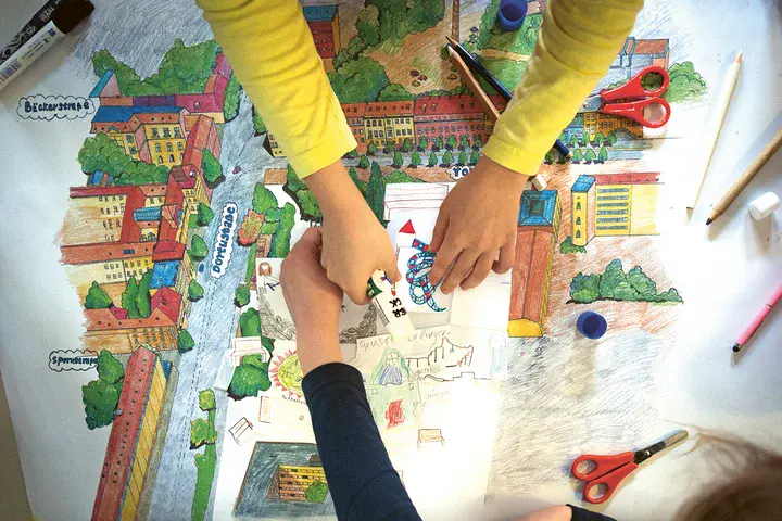 Zwei Paar Kinderhände malen und gestalten auf einem großen Blatt Papier mit bunten Farben und Schere einen Spielplatz