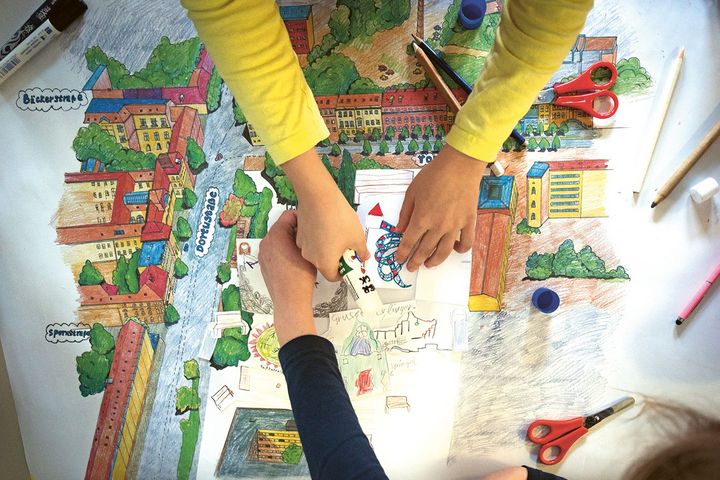 Zwei Paar Kinderhände malen und gestalten auf einem großen Blatt Papier mit bunten Farben und Schere einen Spielplatz