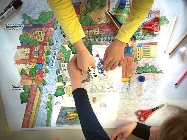 Zwei Paar Kinderhände malen und gestalten auf einem großen Blatt Papier mit bunten Farben und Schere einen Spielplatz