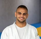 Zwei Judo-Trainer in weißen Anzügen mit schwarzem Gürtel in einer Trainingshalle vor gelb-blauen Sportmatten. 