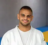 Zwei Judo-Trainer in weißen Anzügen mit schwarzem Gürtel in einer Trainingshalle vor gelb-blauen Sportmatten. 
