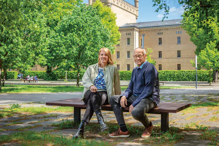 Ein Mann sitzt auf einer Bank. Im Hintergrund ist ein großes ehemaliges Kasernen-Gebäude zu sehen.