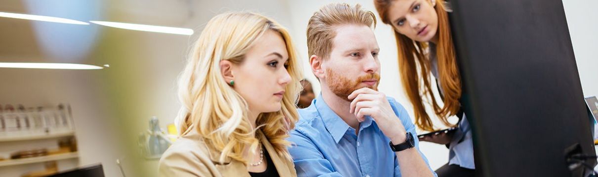 Büro mit computerausgestatteten Arbeitsplatz: im Vordergrund sitzt eine junge, blonde Frau und ein rothaariger Mann, die etwas am Bildschirm anschauen. Eine weitere Frau schaut ebenfalls um den Bildschirm herum.