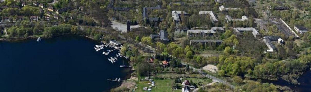 Luftbild von Krampnitz mit einigen Gebäuden und vielen Grünflächen und Bäumen, im Hintergrund ein See.
