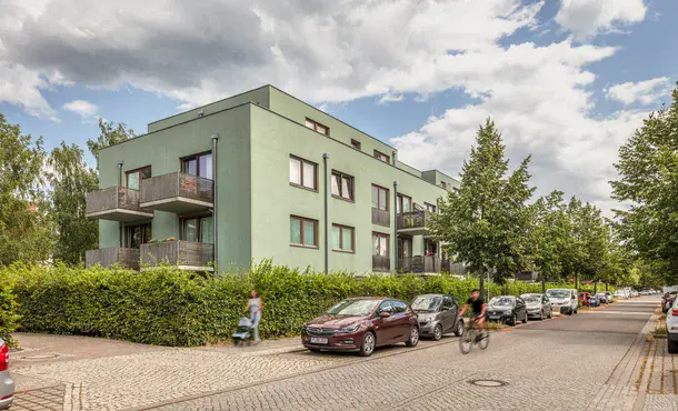 Grünes, mehretagiges Wohnhaus mit grüner Hecke und Bäumen umgeben an einer Straße