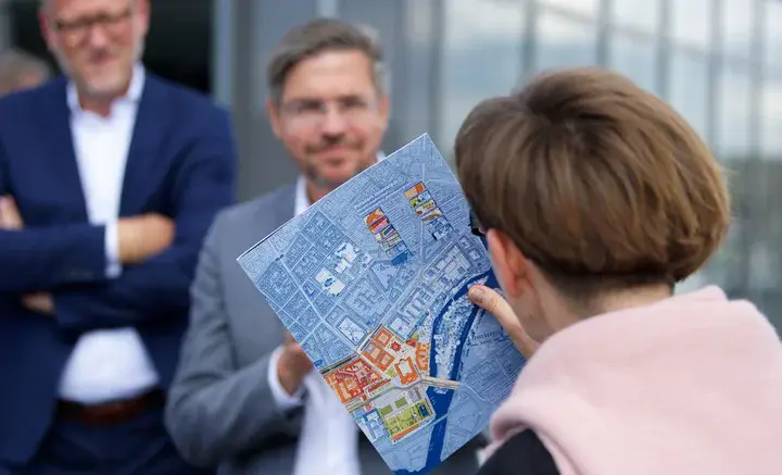 Frau von hinten hält Lageplan in der Hand mit dem sie zwei gegenüber stehenden Männern etwas erläutert