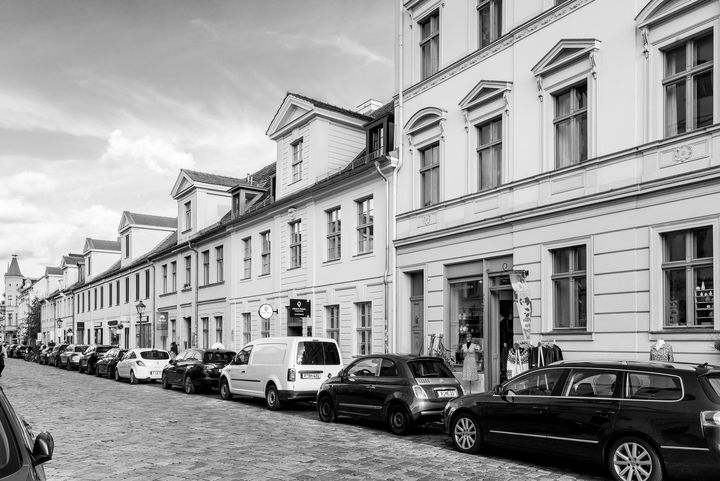 Schwarz-Weiß-Foto einer flachen Häuserzeile an einer Straße mit hintereinander parkenden Autos