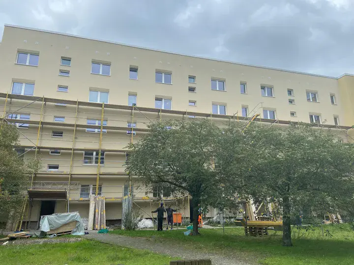 Ansicht einer sanierten Fassade an der die Gerüste abgebaut werden. Im Vordergrund ist eine Grüne Wiese und Bäume zu sehen.
