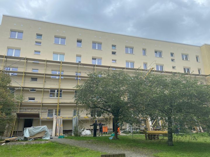 Ansicht einer sanierten Fassade an der die Gerüste abgebaut werden. Im Vordergrund ist eine Grüne Wiese und Bäume zu sehen.