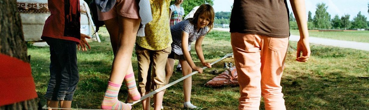 Ein Mädchen balanciert im Park über ein von einem anderen Mädchen gehaltenes Seil, weitere Mädchen stehen dabei.
