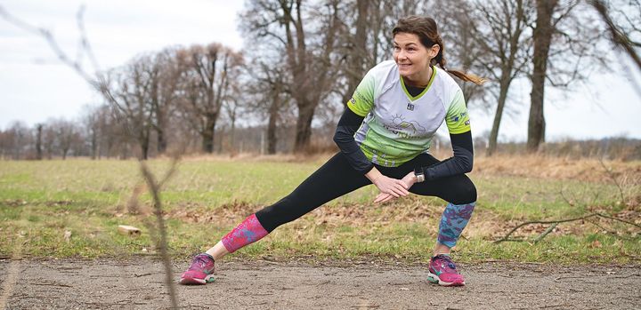 Joggerin dehnt sich auf ihrer Laufstrecke vor einem Feld mit Bäumen im Hintergrund