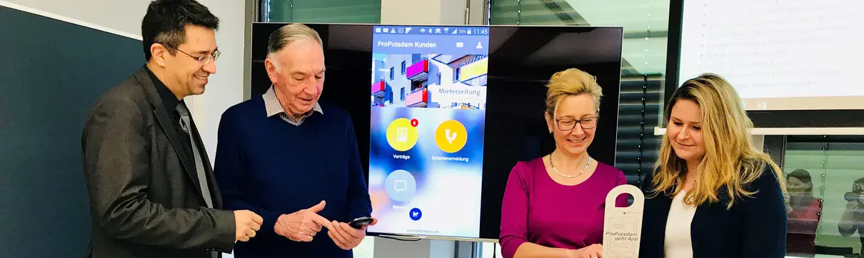Der Geschäftsführer der ProPotsdam und die Geschäftsführerin der Gewoba stehen mit zwei weiteren Personen in einem Raum und schauen auf Handy-Displays.
