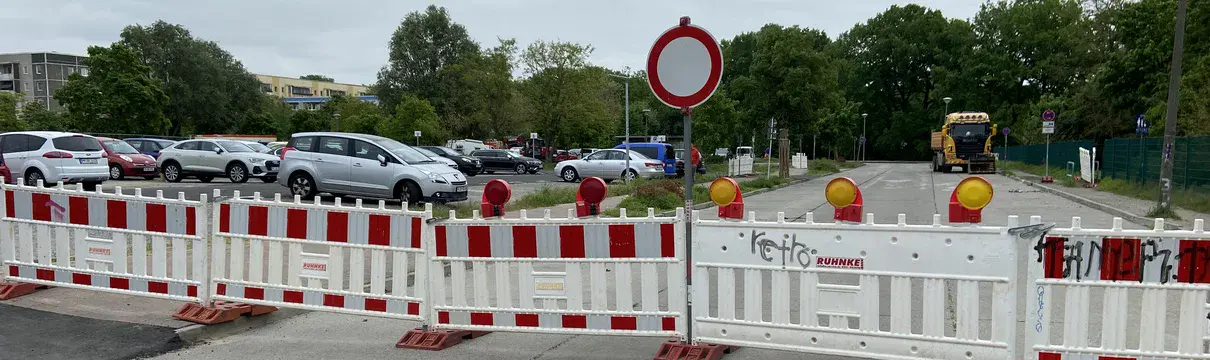 Weiß-Rote Straßenabsperrung mit Straßenschild. Im Hintergrund ist ein Parkplatz zu sehen.