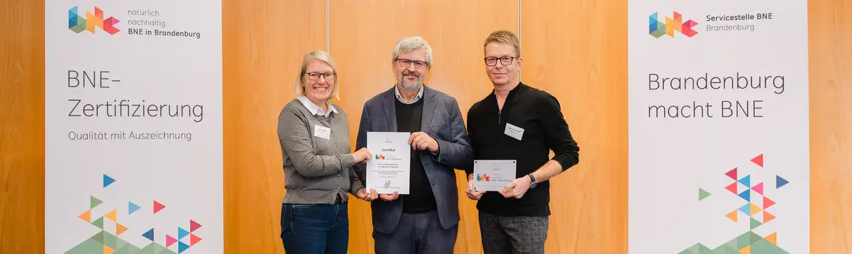 BNE Zertifizierung Grünes Klassenzimmer_(v. l. n. r.) Silke Eggert, Axel Vogel und Olaf Jöllenbeck (c) Christiane Schleifenbaum Drei Menschen stehen vor einer Holzwand und bunten Bannern und halten Urkunden hoch.