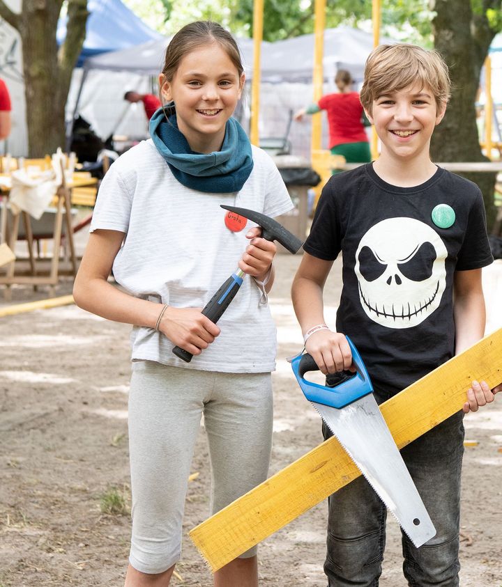 Ein Mädchen mit einem Hammer in der Hand und ein Junge mit einer Säge und einem Holzbrett schauen in die Kamera.