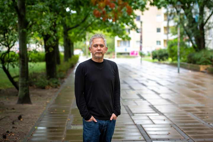 Ein Mann mit Bart steht auf einem kleinen asphaltierten Weg mit Bäumen an den Seiten.