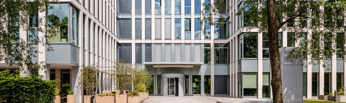 verglastes Bürogebäude vor blauem Himmel, umgeben von hohen, grünen Bäumen