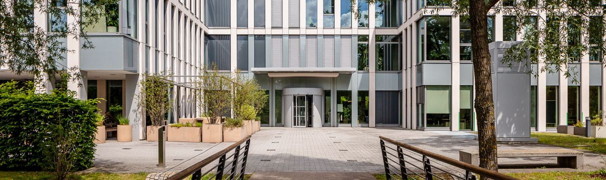 verglastes Bürogebäude vor blauem Himmel, umgeben von hohen, grünen Bäumen