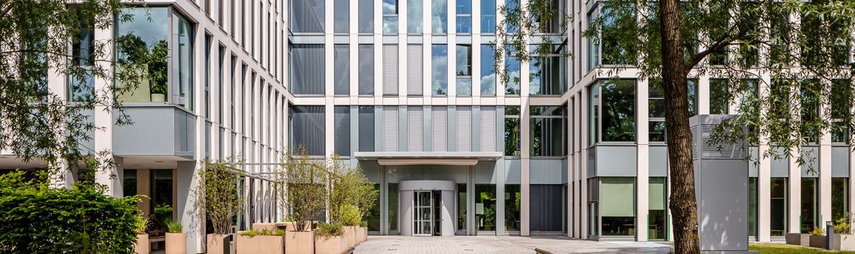 verglastes Bürogebäude vor blauem Himmel, umgeben von hohen, grünen Bäumen