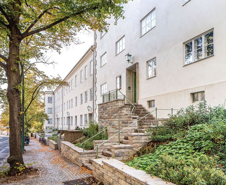 Geschwungener, steinerner Treppenaufgang mit grünem Handlauf zu einem der Eingänge eines hellen Wohngebäudekomplexes an einer Alleestraße