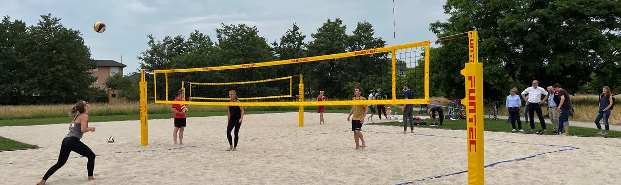 Zu sehen sind Männer und Frauen, die auf einem Sandfeld mit gelbem Netz Beachvolleyball spielen.