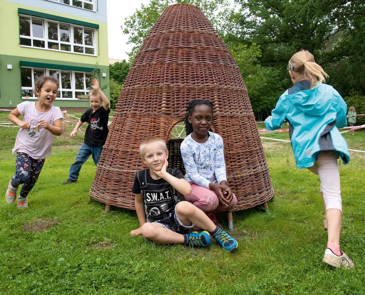 drei Kinder spielen Fange um ein Weidenhäuschen herum, ein Junge und ein Mädchen sitzen lächeln davor im Gras