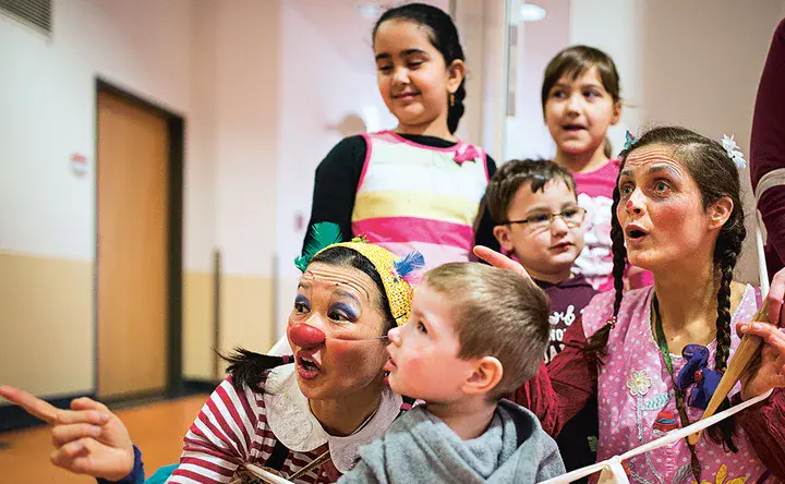 Zwei Clown-Frauen und eine kleine Gruppe Kinder in einem Flur schauen auf etwas hinter dem Betrachter