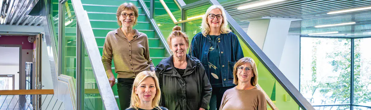Fünf Frauen stehen auf einer grünlichen Treppe in einem modernen Gebäude mit großen Fenstern.